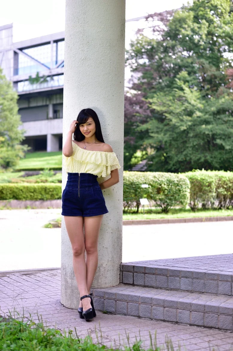 Cô gái mặc áo trễ vai màu vàng và quần short xanh đứng bên hàng cây